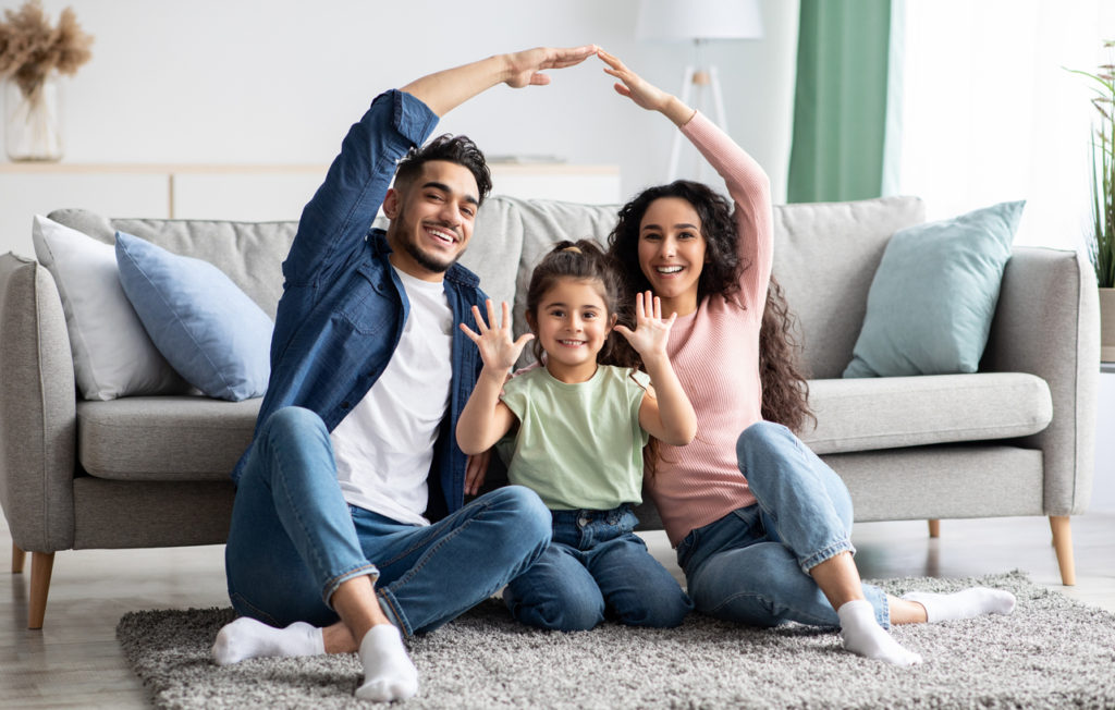 Happy family of three sitting on a rug in a cozy living room, smiling and making hand gestures, representing a pest-free home environment promoted by Rock Pest Control.