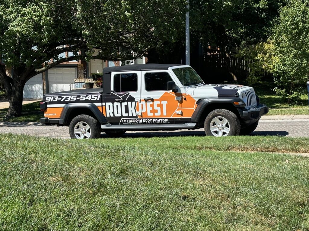 Rock Pest Control branded vehicle parked on a residential street, featuring the phone number 913-735-5451 and the tagline "Leaders in Pest Control," surrounded by green grass and trees.
